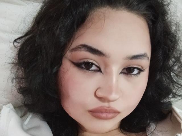 MyrkaHot's face with curly black hair, brown eyes, and winged eyeliner. She has a nose ring and full pink lips, looking serious with a slight pout.