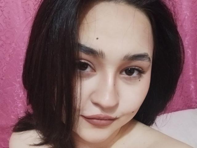 MyrkaHot has straight black hair, brown eyes, and a small mole above her right eyebrow. She's smiling with subtle makeup, showing her cute nose and full, pink lips.