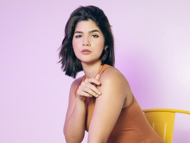 ScarlletZhen tiene el cabello corto y de color marrón oscuro, y lleva una camiseta naranja. Está sentada en una silla amarilla, con la mano cerca de la barbilla, mirando a la cámara..
