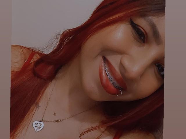 EmmaSeduissante with red hair, black eyeliner, and red lipstick, smiling with a silver tooth, wearing a heart necklace, and a red top, showing her cute face.