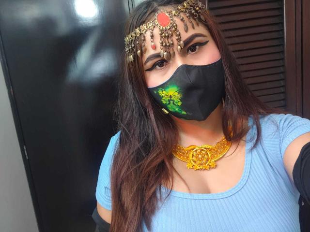 DaliaDubey with dark hair, blue top, black mask with green flower, gold headpiece, red gem, gold necklace, and arm warmers, showing her tits slightly through her top.