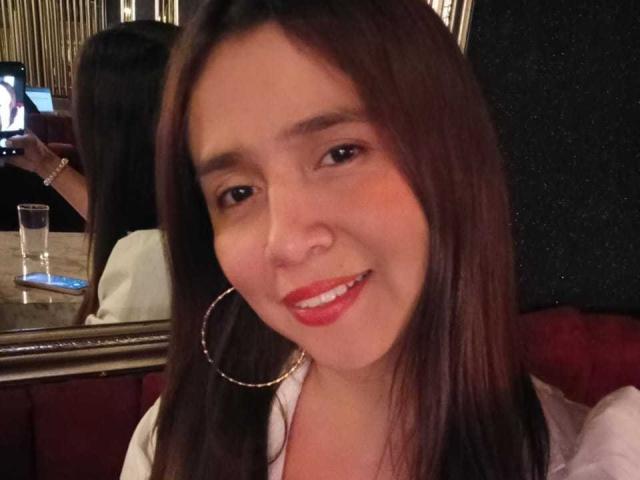 Heidy Wells, mit langen braunen Haaren und rotem Lippenstift, lächelt in die Kamera, trägt große Ohrringe und ein weißes Hemd, in einer schummrigen Bar..