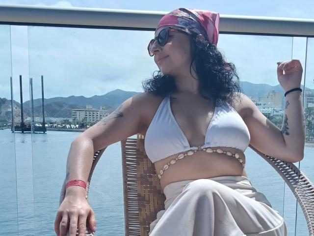AnntonellaWatson with curly black hair, pink headscarf, sunglasses, white bikini top showing her tits, tan skirt, tattoos, wicker chair, holding sunglasses, by water, mountains in background.