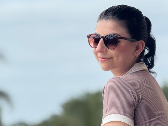 AnntonellaWatson with brown ribbed shirt, white collar, dark brown sunglasses, and ponytail, smiling at camera, showing ass slightly, wearing small pearl earrings, against blurry green and blue background.