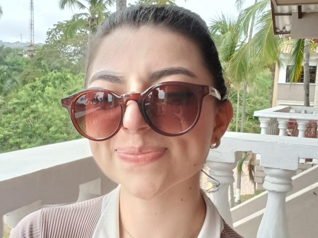 AnntonellaWatson with brown sunglasses, pouty pink lips, and a light brown shirt, smiling slightly, has dark hair in a ponytail, and small gold earrings, standing on a white balcony.