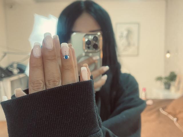 LillyTaylor's hand with long, pink, nude-manicured nails, black sweater, and blue gem on ring finger, holding phone, mirror selfie, blurred face, dark hair, soft light.