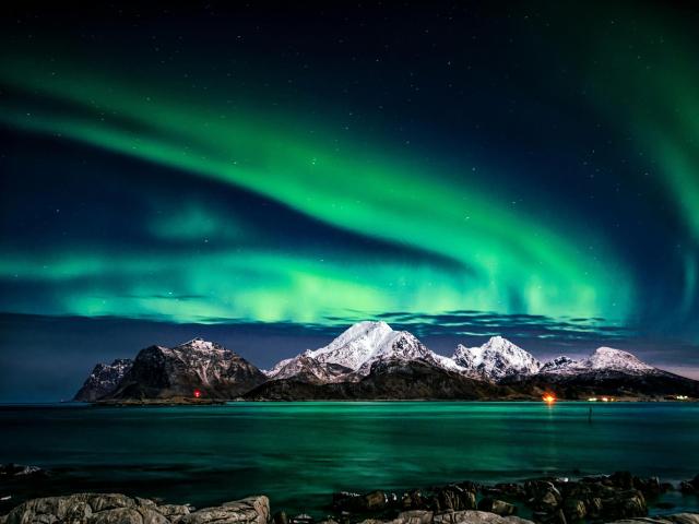A sick-ass night with green Northern Lights dancing over snowy mountains and a calm, dark blue ocean in the foreground. Rocks are in the bottom.