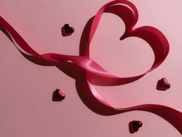 A shiny pink ribbon makes a heart shape, with three small black hearts scattered around on a pink surface. MissSunshine-hot's sexy gift.