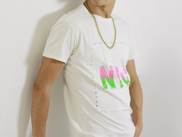 TheRoomSex69 wearing a white t-shirt with pink and green lettering, gold chain, standing against a white wall, showing a muscular arm, and a partially visible face.
