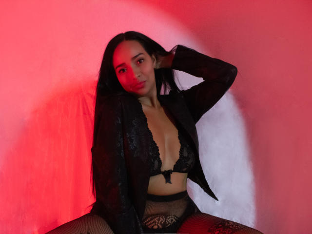 Amber Rousell, con cabello largo y negro, vestida con un sujetador de encaje negro y una chaqueta transparente, con luces rojas y blancas sobre ella, con la mano en el cabello, sentada con las piernas separadas, mostrando pantalones de malla de pescado..
