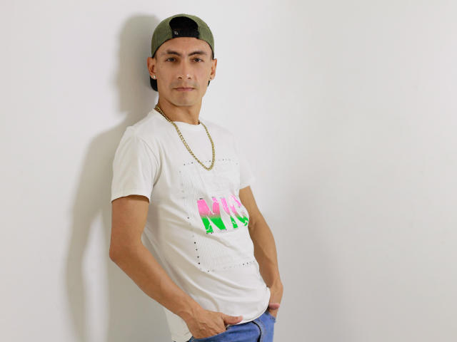 TheRoomSex69 is a slim dude with tan skin, wearing a green cap, white tee, gold chain, and blue jeans, standing against a white wall, hands in pockets.