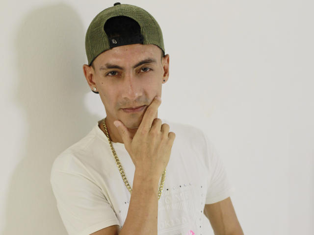 TheRoomSex69, wearing a green backward cap, white t-shirt, and gold chain, with hand on cheek, smiling subtly. Medium skin, brown eyes, small earrings, white wall in background.