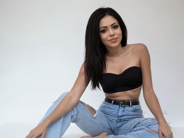 KittyMissyX with black strapless top, showing her small tits, and ripped blue jeans, sitting with legs spread, long black hair, and a slight smile, barefoot.