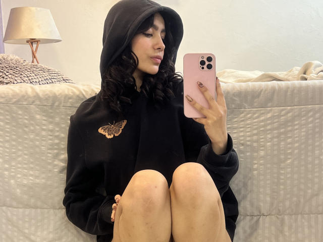 AitanaJohns with curly black hair, wearing a black hoodie with a butterfly emblem, sits on a bed, holding a pink iPhone, taking a selfie, knees up.