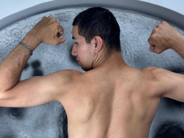 MartinBlos, shirtless with a muscular back, flexing both arms, showing defined triceps and biceps, wearing a silver bracelet, short dark hair, slightly tanned skin, looking to the side.