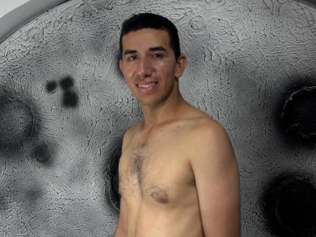 MartinBlos, shirtless, smiling, short dark hair, light skin, small chest with nipples, slight chest hair, standing in front of a textured, frosted glass surface.