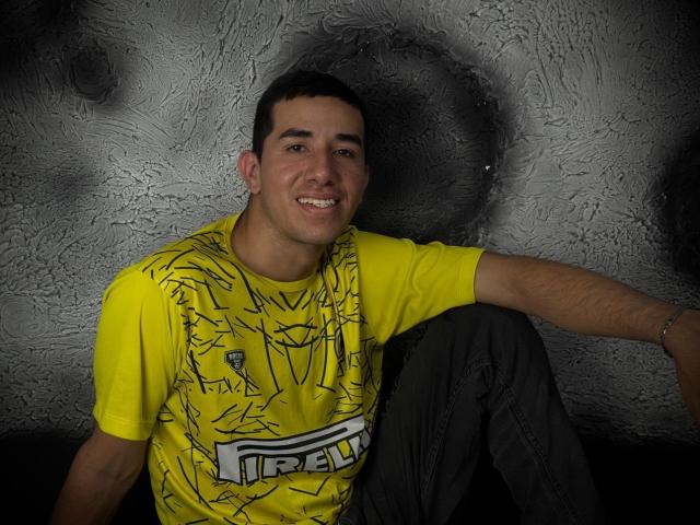 MartinBlos, with short dark hair and a smile, wearing a yellow t-shirt with black line art and gray pants, sitting with one arm on a dark surface.