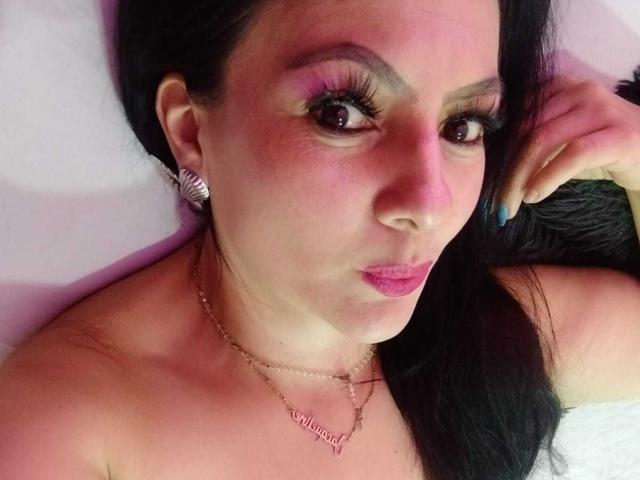 JakyKidman with pink lipstick, purple eyeshadow, long black hair, butterfly earrings, gold necklace, pouty lips, blue nail, lying down, pinkish skin, looking at camera, slight blush.