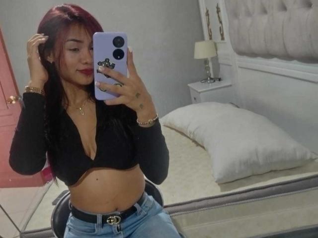 SashaHans, con cabello rojo, un top corto negro que deja ver sus pechos, y jeans azules, tomando una selfie frente a un espejo, sosteniendo un teléfono morado, en un dormitorio..