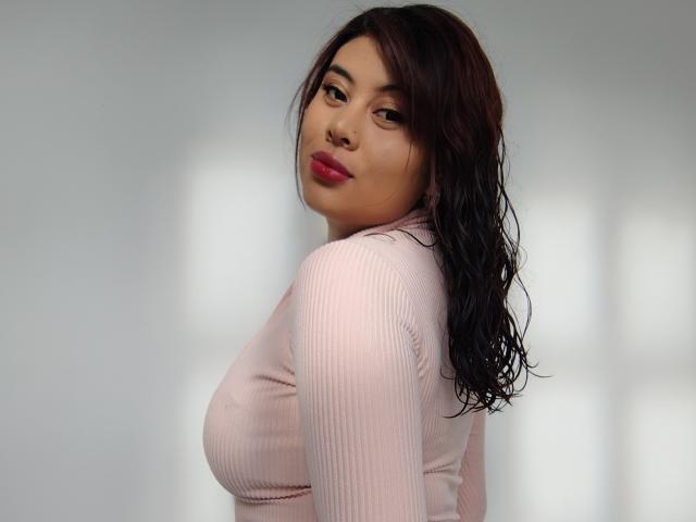 NoemiThornee met nat zwart haar, een roze geribbelde top en rode lippenstift, kijkt over haar schouder en laat haar grote borsten zien. Zacht licht op haar gezicht, een subtiele glimlach..