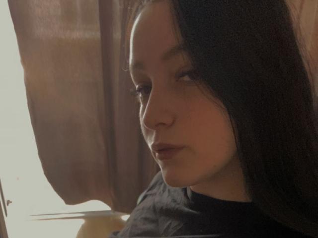 LeahBrooks has dark hair, light skin, and is looking to the side with a slightly pouty expression, wearing a black shirt, in a dimly lit room.