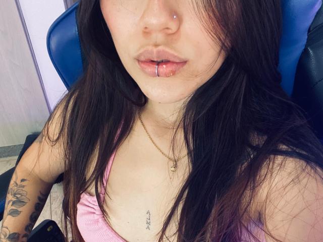 SophiaMarx with black hair, nose piercing, lip ring, pink top, tattooed arm, necklace, and visible cleavage, sitting in a blue chair. Her pussy is not visible.