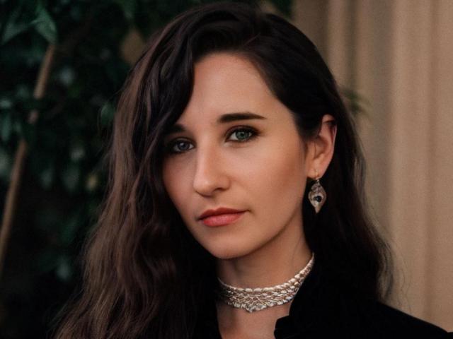NetaRose ha i capelli castano ondulati, gli occhi verdi e un'espressione seria con le labbra rosate. Indossa una maglia nera, una collana di perle e orecchini pendenti. Il suo seno è coperto..