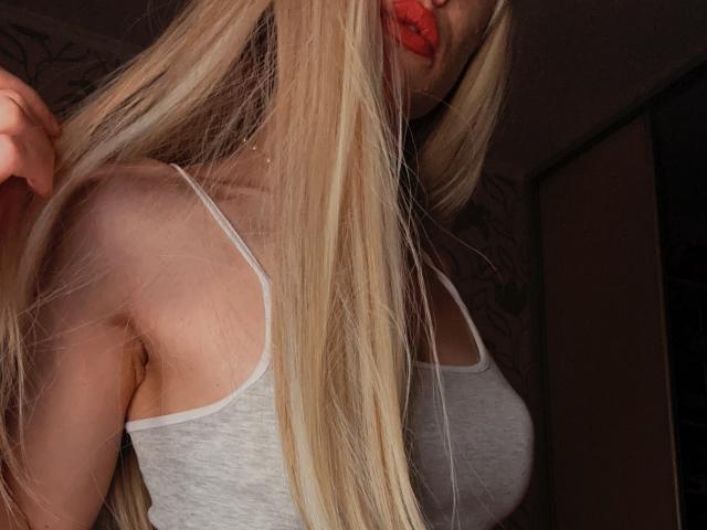 Kirana with long blonde hair, red lips, wearing a grey tank top, showing her small tits and bare shoulder. She's holding her hair, looking down. Dimly lit.