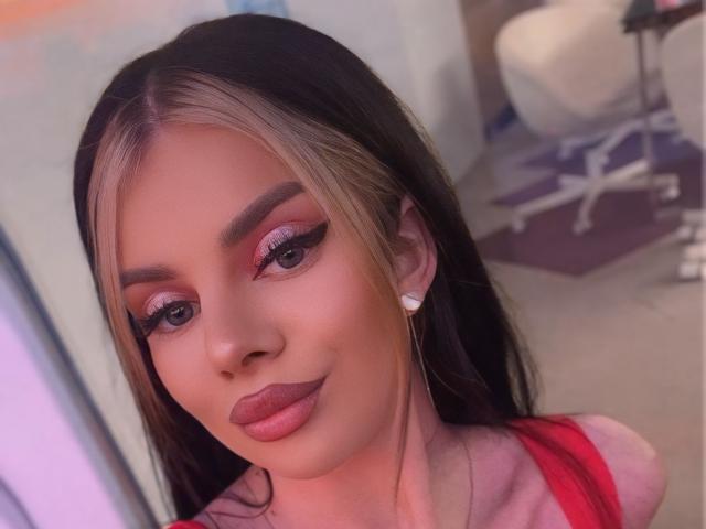 IvyRaven with dark brown hair, blonde highlights, pink eyeshadow, thick eyeliner, red lipstick, and white earrings, smiling at the camera, wearing a red top, in a bedroom.