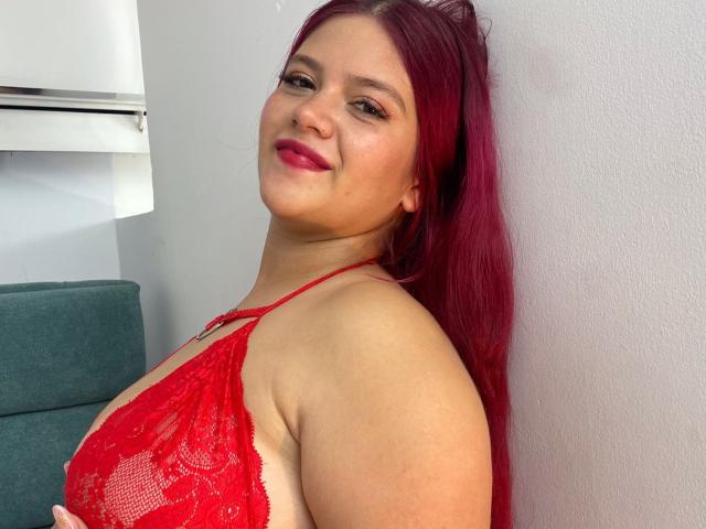 RubyMartin with red lace bra, showing her tits, has pink hair, red lipstick, and a slight smile, leaning against a white wall, looking at the camera.