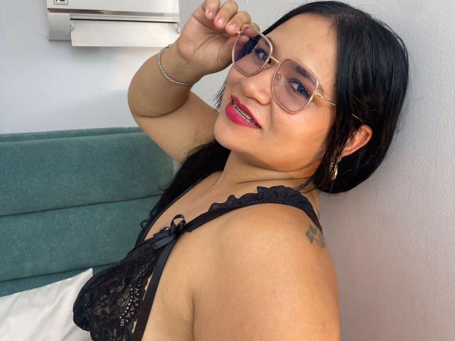 EvaJohns with glasses, black lace bra, red lipstick, black hair, tattoo on shoulder, smiling, adjusting glasses, green headboard, white wall, silver bracelet, looking at camera.