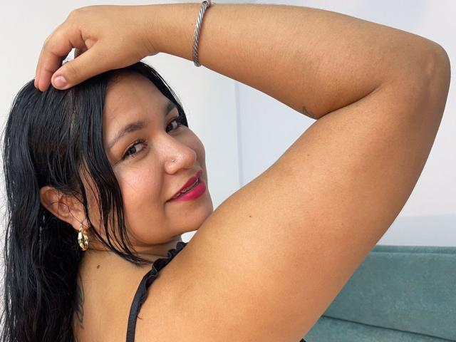 EvaJohns with dark hair, tan skin, and pink lipstick, arms raised, showing her armpit, wearing gold earrings, black top, and silver bracelet, smiling slightly.