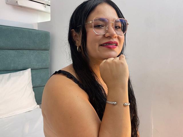 EvaJohns with long black hair, glasses, pink lipstick, black bra, and bracelet, resting chin on hand, smiling slightly. Her tits are small and she's cute.