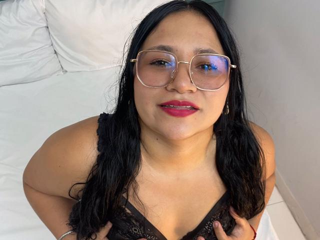 EvaJohns with black lace top, holding her tits, wearing glasses, red lipstick, long black hair, and a slight smile, sitting on a white bed.