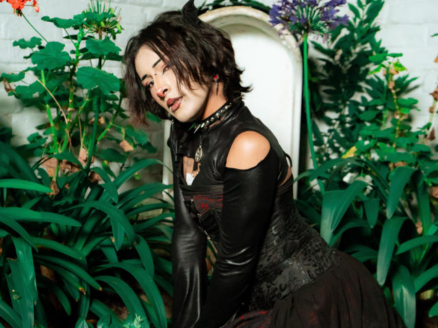 NoemiSky with wavy black hair, leather top, and black skirt, leaning on green plants, wearing a spiked choker, looking seductively at the camera, showing her small tits.