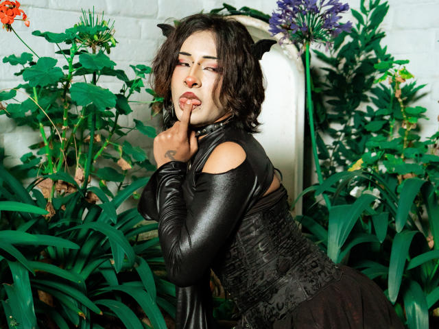 NoemiSky con orejas de gato, mangas de cuero negro y un corpiño, con un dedo en la boca, luciendo sexy, rodeada de plantas verdes, vestida con un vestido negro, mostrando su trasero..