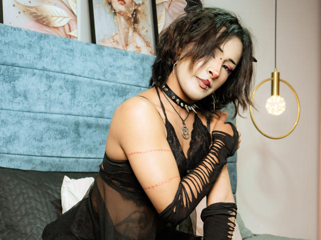 NoemiSky with dark, messy hair, black lace lingerie, and cut-out gloves, showing off her tits and pussy scars, wearing a spiked choker, sitting on a bed.