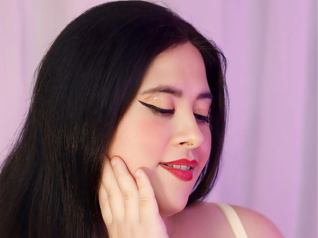 SarahMat with black hair, pinkish skin, red lipstick, black eyeliner, and a nose ring, smiling with eyes closed, touching her cheek with her right hand.