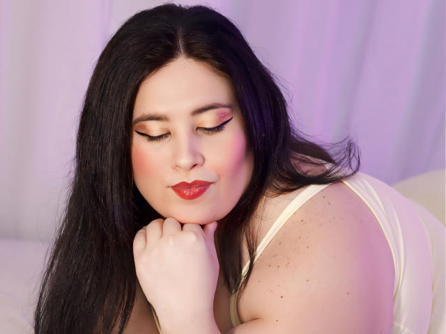 SarahMat with black hair, pink eyelids, red lipstick, white tank top, lying down, hand on chin, eyes closed, soft pink and white background. Boobs partially visible.