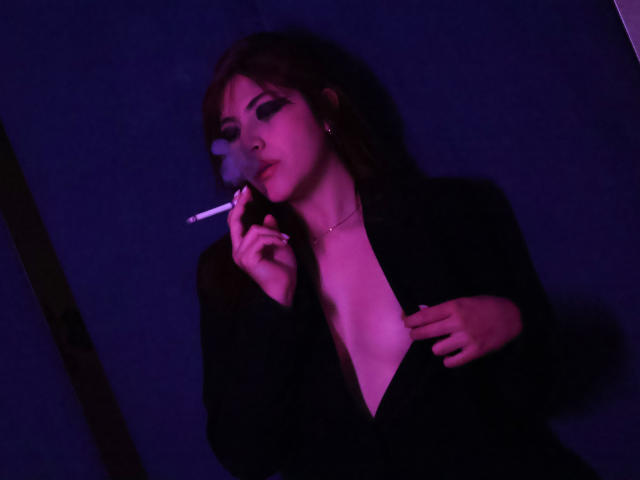 SpencerLe with dark hair, smoking a cigarette, wearing a black blazer, open chest, purple lighting, holding blazer with right hand, left hand holding cigarette, looking down.