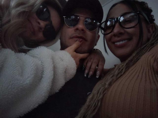 ThreesomeBoom avec une femme blonde, ThreesomeBoom avec un bonnet et des lunettes de soleil, ThreesomeBoom avec des lunettes et un sourire. La main de la femme blonde sur le visage de ThreesomeBoom. ThreesomeBoom au centre, portant une chemise sombre..