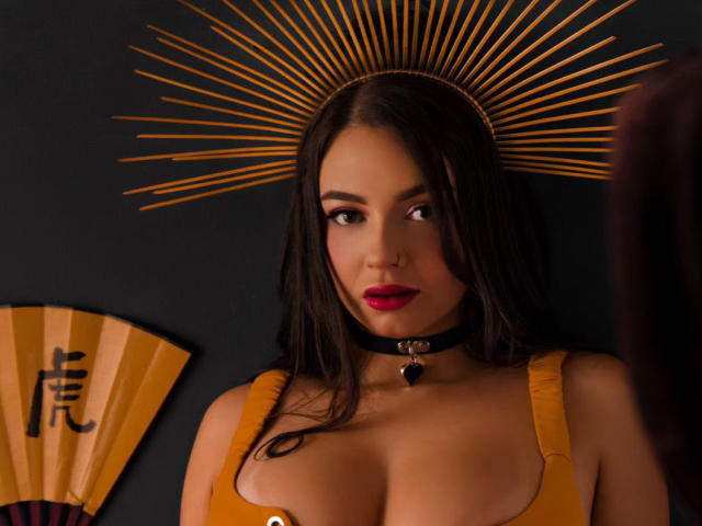 HotSaraSmith69 con capelli scuri, labbra rosse e una maglia gialla, indossa una collana nera con un cuore, un copricapo a forma di sole e guarda di lato, mostrando il suo seno prosperoso..