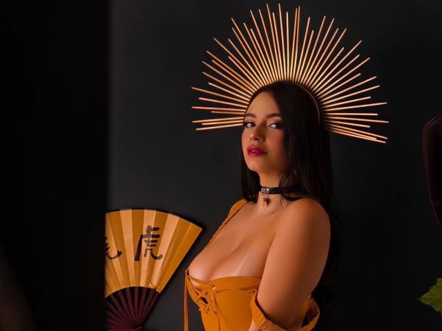 HotSaraSmith69 con grandi seni, corpetto arancione, collana nera e copricapo a forma di alone solare, che guarda di lato, capelli scuri, rossetto rosso, ventaglio in mano sullo sfondo..