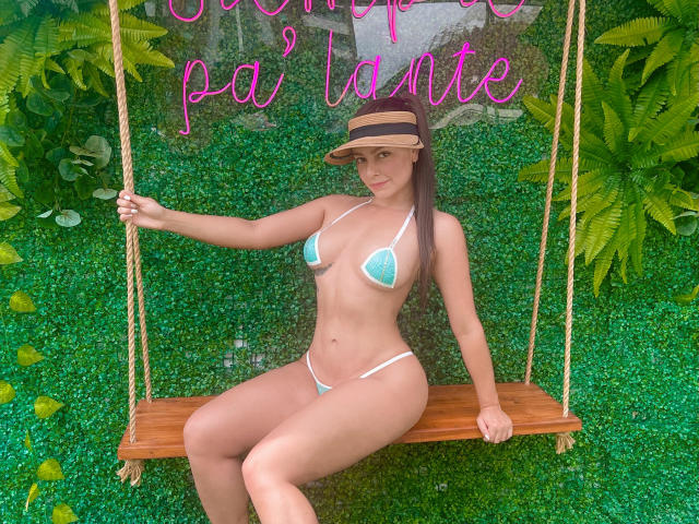 HotSaraSmith69, con la pelle abbronzata, indossa un bikini color turchese, una visiera marrone e ha i capelli castani, seduta su un'altalena di legno, tenendo una corda, con erba verde e felci sullo sfondo..