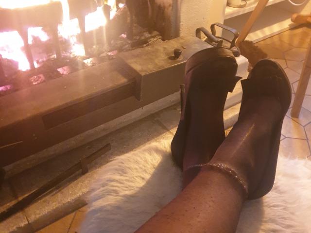 LadyNocturne's legs with dark purple high-heeled shoes, ankles adorned with sparkly ankle bracelets, resting on a white fur rug in front of a lit fireplace.