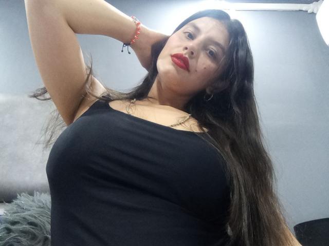 MadissonFontaine with long brown hair, red lipstick, black tank top, and red bracelet, showing off her big tits and raised arm. She's looking down at the camera.