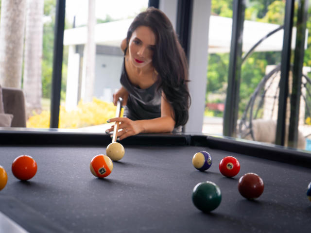 EvelinLuna with long dark hair, red lipstick, and gray top, leaning over black pool table, aiming cue stick at orange ball, surrounded by colorful pool balls.