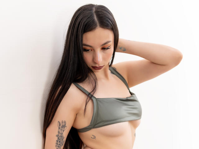 CamilleRios has long black hair, olive skin, small breasts, green crop top, tattoos on arm and side, dark lipstick, and a hand in her hair, looking down.