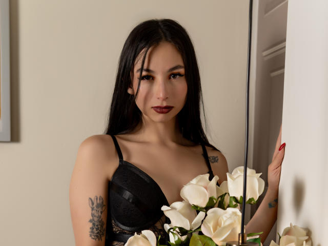 CamilleRios with long black hair, wearing a black lace bra, holding white roses, has tattoos on her right arm, red lipstick, and red nails, standing near a white wall.
