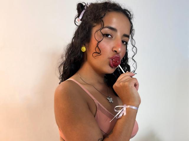 LunnaBenson with curly black hair, pink bra, yellow earrings, sucking a red lollipop, holding a lipstick, wearing a white ribbon bracelet, looking at the camera.